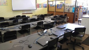 The view from the back-left corner of the computer lab shows the traditional layout of desks facing the front projector. The entry door is on the right-hand side.